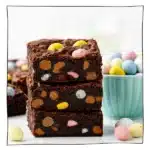 Eggless Cadbury Mini Egg Brownies stacked on a white surface with colorful candy pieces and milk in background