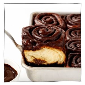 Close-up of a vegan chocolate cinnamon roll with gooey chocolate glaze, lifted from a white baking pan and showing its fluffy egg-free interior.